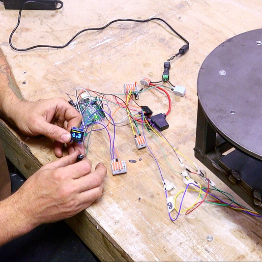 A man working at a wooden table with an electronic circuit board, wires, and a round metal platform on a workbench.