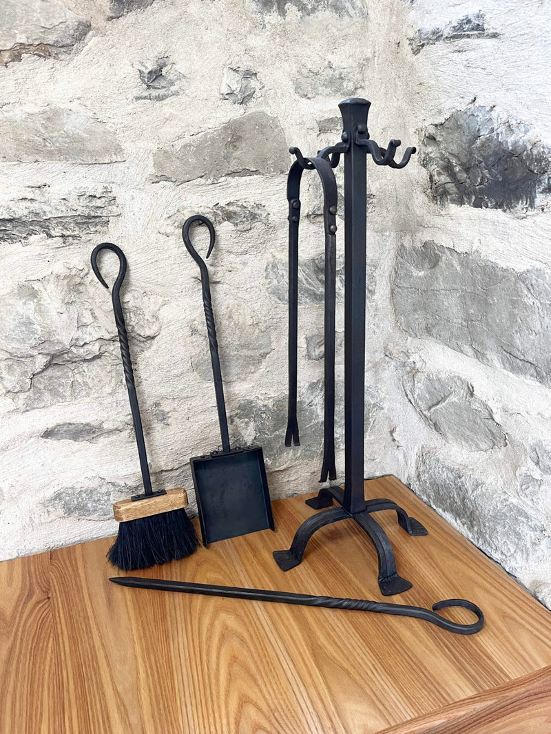 Set of black wrought iron fireplace tools including a brush, shovel, and tongs on a wooden surface with a stone wall background.