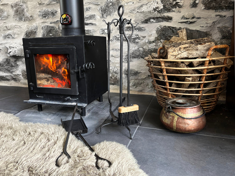 Wood-burning stove with firewood basket and tools against a stone wall.