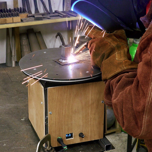 A person in a welding helmet and leather protective gear welds metal on a wooden table in a workshop, with bright sparks flying. The welding helmet has a blue glow, and the background shows tools and equipment on shelves.